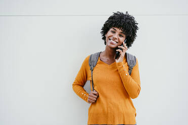 Smiling woman talking on mobile phone in front of white wall - EBBF06699