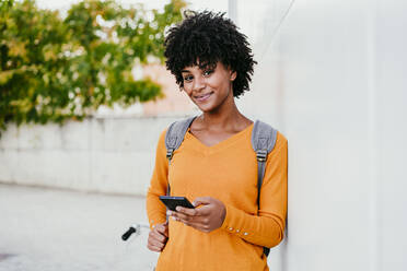 Smiling young woman with mobile phone by wall - EBBF06701