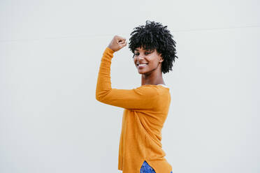 Smiling young woman flexing muscle near white wall - EBBF06704