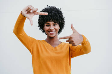 Smiling young woman gesturing picture frame in front of wall - EBBF06707