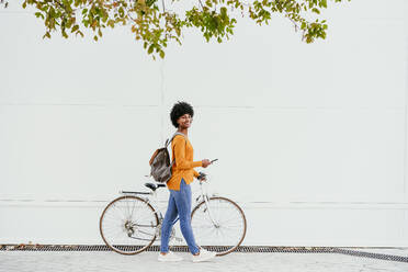 Smiling woman with backpack and smart phone wheeling bicycle by wall - EBBF06712