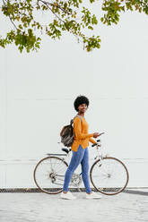 Young woman holding smart phone walking with bicycle by wall - EBBF06713