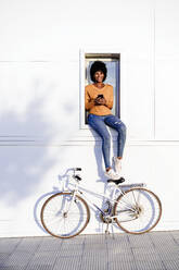 Young woman with bicycle with mobile phone sitting on window - EBBF06719