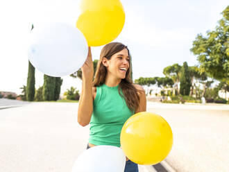 Lächelnde Frau mit gelben und weißen Luftballons, die sich am Fußweg erfreut - AMRF00096