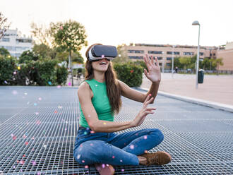 Glückliche Frau mit VR-Brille, die auf einem Metallfußweg sitzt - AMRF00104