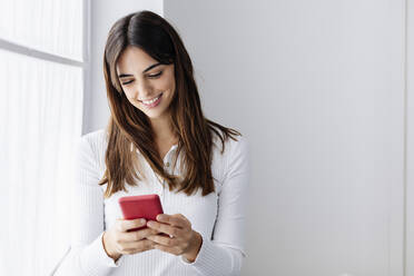 Lächelnde Frau, die ihr Smartphone am Fenster zu Hause benutzt - XLGF03110