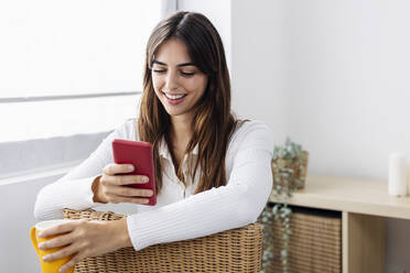 Smiling woman holding coffee cup using smart phone on chair at home - XLGF03117