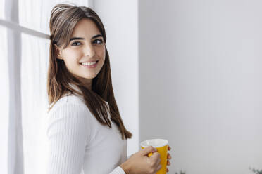 Smiling woman holding coffee cup leaning on window - XLGF03119