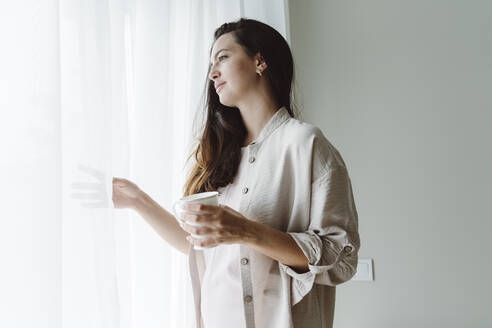 Nachdenkliche Frau, die eine Kaffeetasse hält und durch ein Fenster zu Hause schaut - OSF01078