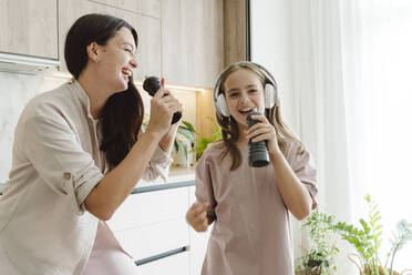 Happy girl with mother singing into pepper mills and enjoying in kitchen - OSF01080