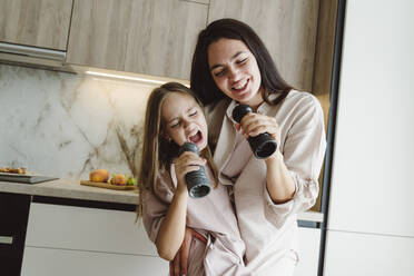 Happy mother and daughter singing into pepper mills at home - OSF01081