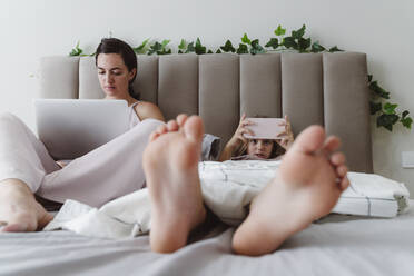 Woman with laptop sitting by girl using smart phone lying on bed at home - OSF01083