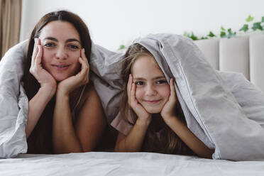 Smiling woman and daughter with head in hands under blanket lying on bed - OSF01086