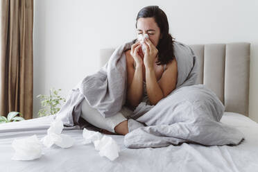 Ill woman with eyes closed blowing nose in tissue on bed at home - OSF01097