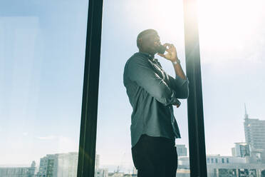 Geschäftsmann im Gespräch am Handy in einem modernen Büro mit Blick auf die Stadt und Sonnenlicht. Afrikanischer Mann im Gespräch am Telefon, während er an einem Fenster im Büro steht. - JLPSF13830