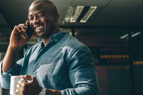 Lächelnder junger Mann mit Kaffeetasse in der Hand, der in einem modernen Büro mit dem Handy telefoniert. Glücklicher Geschäftsmann, der im Büro telefoniert. - JLPSF13832