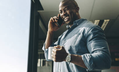 Happy young businessman with coffee in hand talking on mobile phone office. Businessman smiling while talking on phone. - JLPSF13833