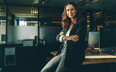 Mature businesswoman sitting on her desk. Confident female entrepreneur with her arms crossed looking at camera. - JLPSF13840