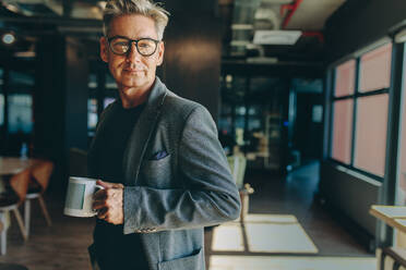 Ein Geschäftsmann steht im Büro und hält eine Kaffeetasse. Ein Unternehmer macht eine Kaffeepause im Büro und schaut in die Kamera. - JLPSF13850