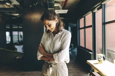 Woman standing in office with arms crossed looking down and smiling. Businesswoman looking happy in office. - JLPSF13857