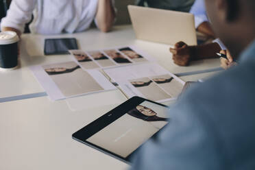 Eine Gruppe von Geschäftsleuten, die Material für ein Projekt auswählen, sitzt um einen Tisch mit einem Foto auf einem digitalen Tablet und Fotoabzügen. - JLPSF13860