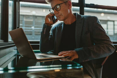 Businessman talking over phone while sitting in front of laptop. Man sitting with a laptop computer talking over phone. - JLPSF13877