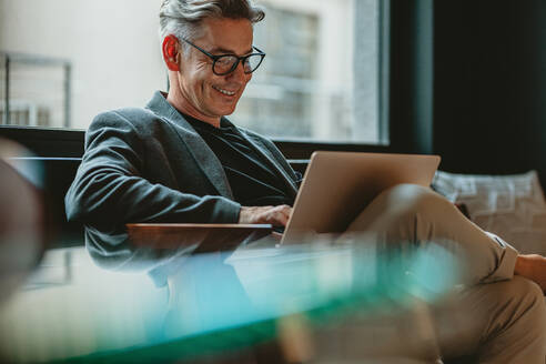 Smiling businessman sitting in office lobby working on laptop. Male business professional working in office lobby. - JLPSF13881