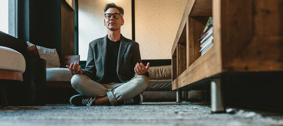 Mature businessman doing yoga meditation in office lounge. Male business executive meditating in lotus pose on the floor in the office. - JLPSF13890
