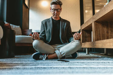 Geschäftsmann sitzt auf dem Boden und meditiert in Yogapose. Älterer Geschäftsmann macht Entspannungsyoga in der Bürolounge. - JLPSF13891
