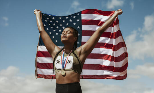 Weibliche Athletin mit Medaille, die mit geschlossenen Augen die amerikanische Flagge hält. Stolze junge Frau in Sportkleidung mit der Flagge der Vereinigten Staaten von Amerika. - JLPSF13916