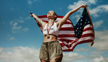 Sportler, der seinen Sieg mit der US-Nationalflagge gegen den Himmel feiert. Läuferin mit Medaillen, die einen Sieg feiert und die amerikanische Flagge hält. - JLPSF13917