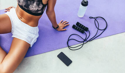 Ausgeschnittene Aufnahme einer Frau in Fitnesskleidung, die sich nach dem Training auf einer Yogamatte mit Handy, Wasserflasche und Springseil neben sich entspannt. - JLPSF13937