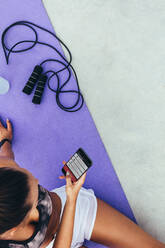 Draufsicht auf eine junge Frau, die auf einer Trainingsmatte sitzt und eine Fitness-App auf ihrem Handy benutzt. Eine Frau benutzt eine Trainings-App zum Trainieren. - JLPSF13940