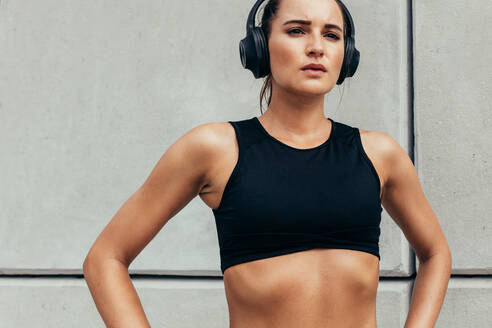 Confident sportswoman standing with her hands on hips and looking away. Fitness girl with headphones on taking break after workout. - JLPSF13948
