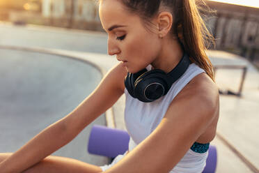 Fitness female sitting with headphones around her neck after exercising. Woman resting after workout outdoors. - JLPSF13957