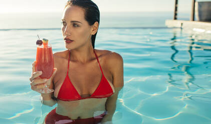 Schöne Frau im Schwimmbad mit Cocktail. Frau im Bikini steht in einem Pool mit einem Getränk und schaut weg. - JLPSF13978