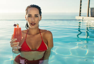 Schöne Frau im Bikini steht in einem Pool mit einem Getränk. Weibliches Model im Schwimmbad mit Cocktail in der Hand schaut in die Kamera. - JLPSF13979