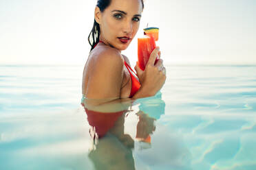 Junge Frau hält einen Cocktail in der Hand, während sie in einem Infinity-Pool steht. Frau im Bikini trinkt einen Fruchtsaft in einem Schwimmbad an einem Sommertag. - JLPSF13981