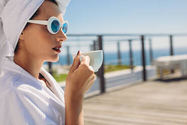 Beautiful woman sitting by pool having coffee. Female model wearing bathrobe and sunglasses relaxing at the poolside. - JLPSF13985