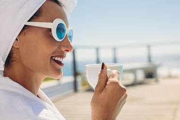 Nahaufnahme einer schönen jungen Frau am Pool, die einen Kaffee trinkt. Weibliches Model im Bademantel und mit Sonnenbrille entspannt sich im Resort. - JLPSF13986