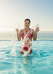 Woman splashing water while standing in a infinity pool. Female in bikini playing in the swimming pool on a summer day. - JLPSF13993