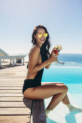 Schöne Frau im Bikini am Pool sitzend mit einem Drink. Frau mit Sonnenbrille entspannt am Rande eines Swimmingpools mit Cocktail in die Kamera schauend und lächelnd. - JLPSF13997