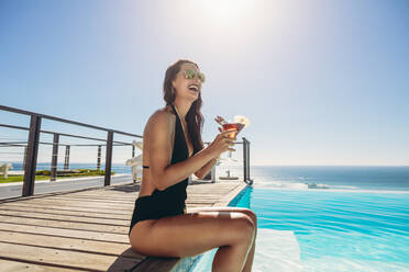 Schöne Frau in Bikini und Sonnenbrille mit einem Getränk am Pool. Weibliches Model genießt einen Sommertag am Swimmingpool des Resorts. - JLPSF13999
