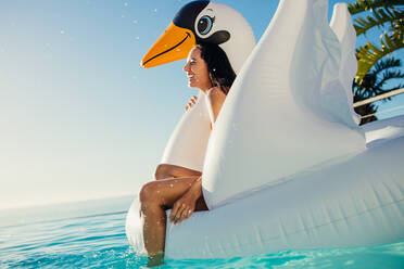 Fröhliche junge Frau lächelt, während sie auf einem aufblasbaren Schwan schwimmt. Frau hat Spaß auf schwimmendem Spielzeug im Schwimmbad. - JLPSF14039