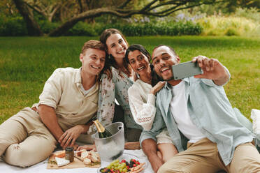 Young man taking selfie with friends at the picnic. Multi-ethnic group of young men and women posing for a selfie at park. - JLPSF14074