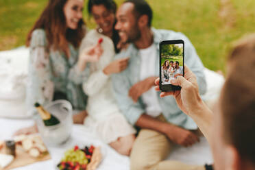 Close-up of a man taking a video of his friends at picnic. Man recording his friends at the park. Focus on mobile phone. - JLPSF14077