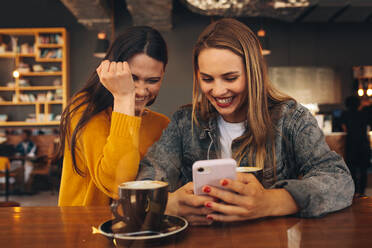 Happy female friends at a cafe using smart phone with a cup of coffee on the table. Women friends looking at a mobile phone and laughing. - JLPSF14081