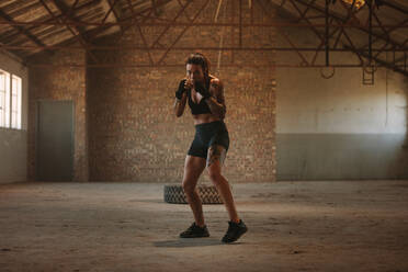 Woman doing shadow boxing exercise in empty factory shade. Woman in sportswear practicing her punches at an abandoned warehouse. - JLPSF14107