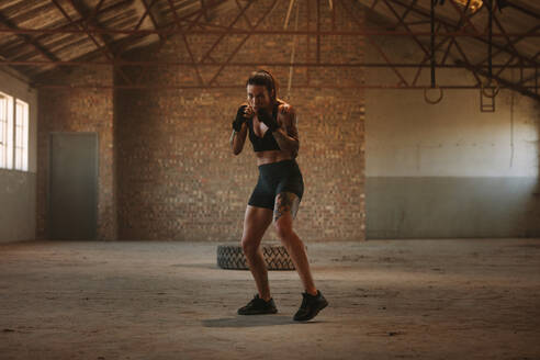 Frau beim Schattenboxen im Schatten einer leeren Fabrik. Frau in Sportkleidung übt ihre Schläge in einer verlassenen Lagerhalle. - JLPSF14107