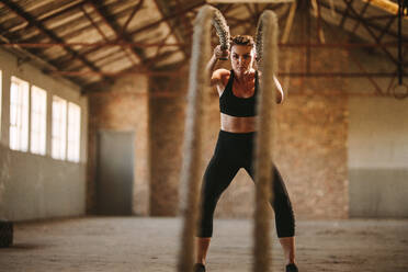 Strong woman working out with battle rope inside abandoned factory shade. Fitness woman exercising with battle rope at cross training gym inside old warehouse. - JLPSF14111